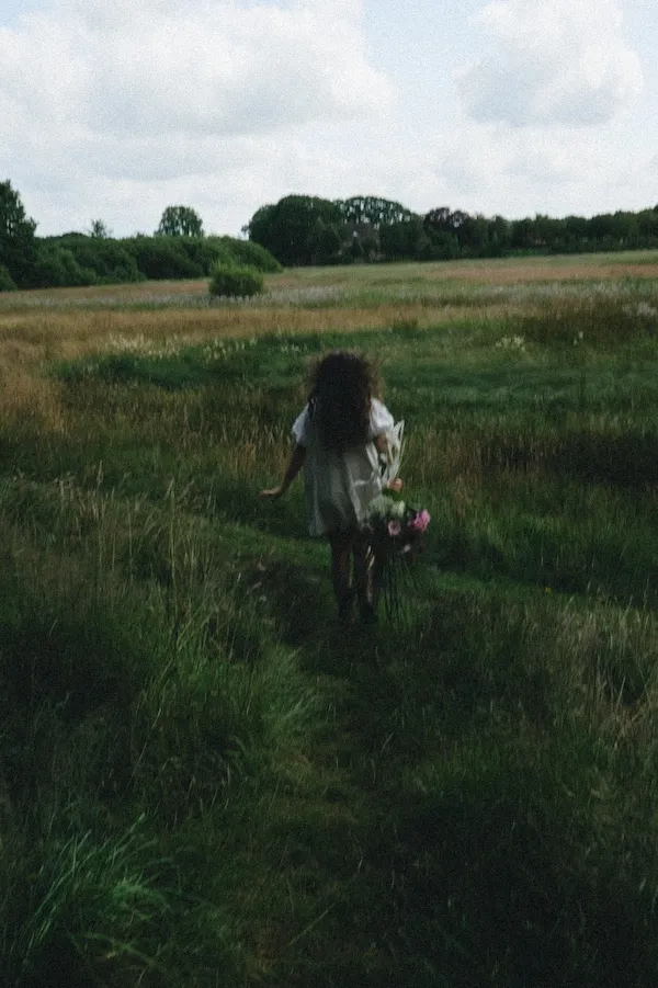 woman in grassy field holding flowers walking away from camera